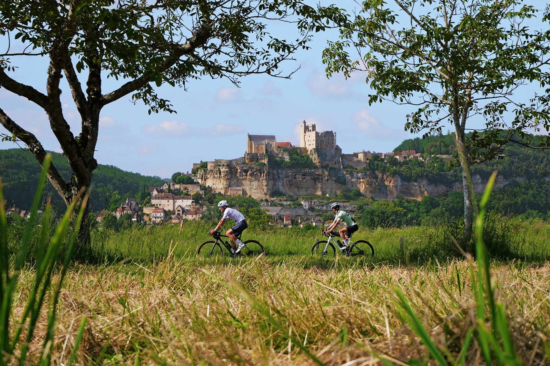 Deux cyclistes roulent dans la campagne avec en arrière plan un village pittoresque surmonté d'un château