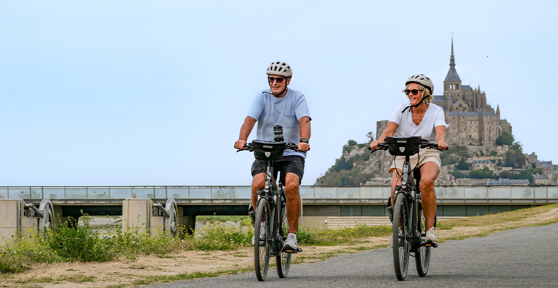 Un couple de cycliste roule sur une petite route avec en fond le Mont saint Michel