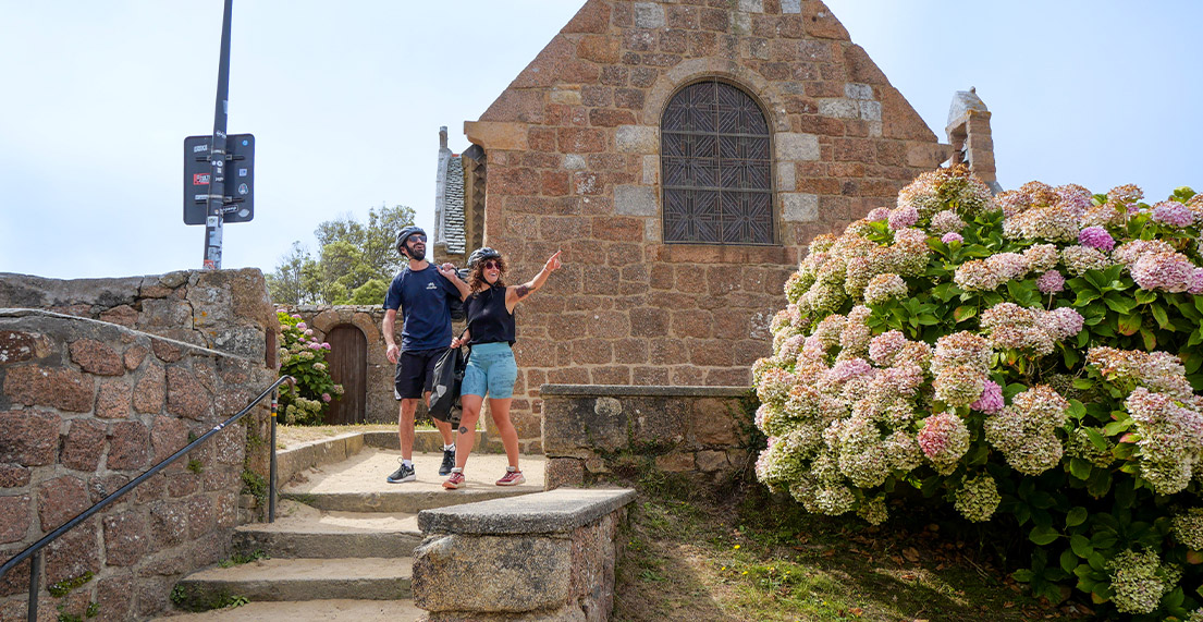 Un couple de cycliste visite et passe devant une eglise rose entoure d hortensias