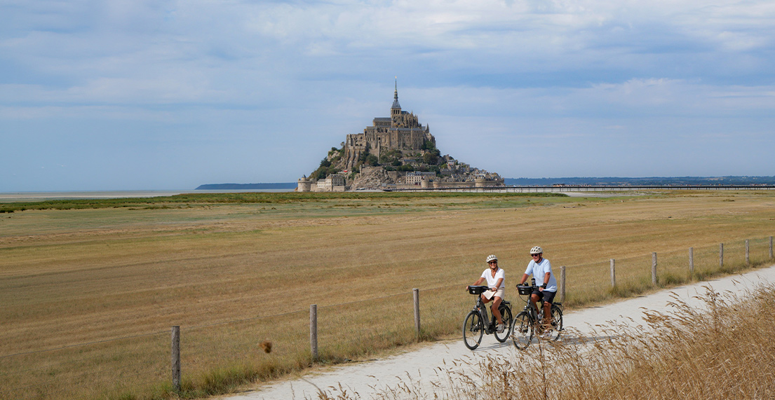 Un couple de cyclistes en vae roule sur un chemin blanc avec le Mont saint Michel qui se dresse derriere eux
