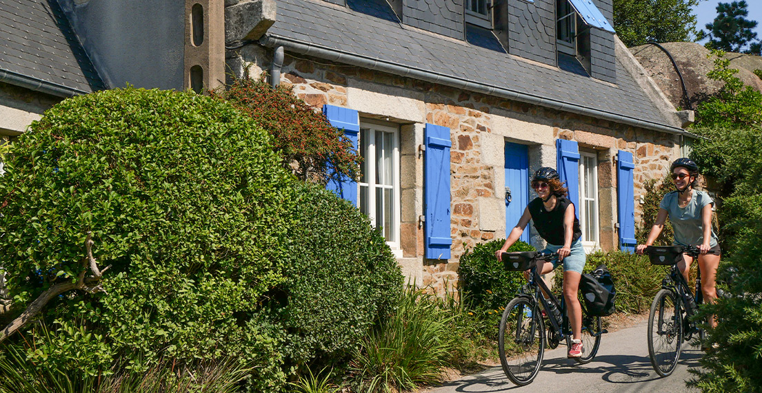 Deux femme sur leur velo roulent devant une maison de pierres roses