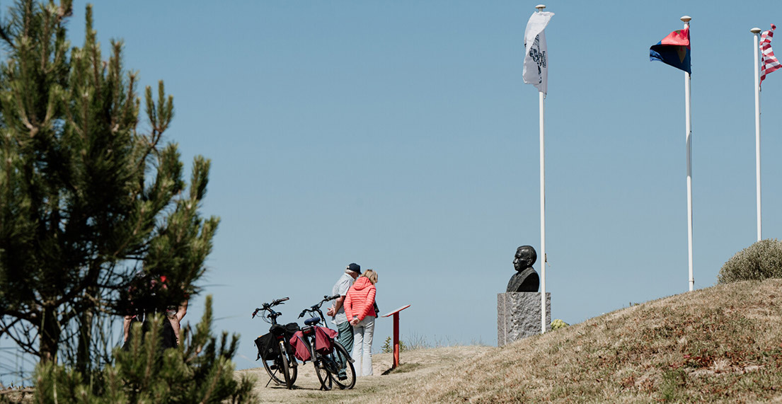 Un couple de cycliste lit un panneau d'information devant une statue et des drapeaux