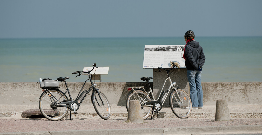 Une cycliste fait un arret devant un panneau informatif devant une des plages du debarquement