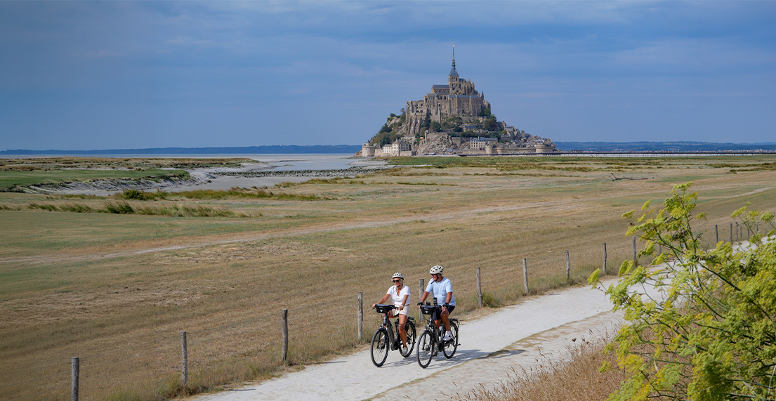 Un couple de cyclaistes roule sur un chemin blanc avec le mont saint michel qui se dresse derriere eux