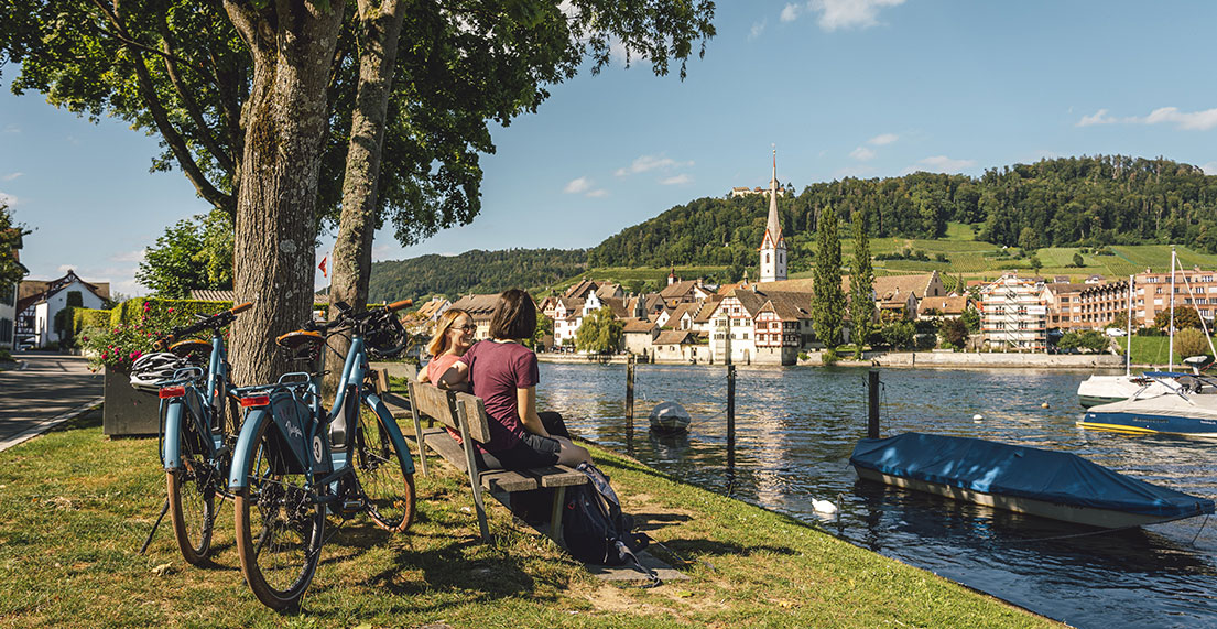 Deux cyclsites assises sur un banc devant le rhin et une ville typique de suisse