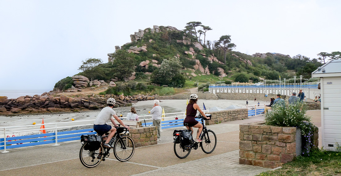 Un couple de cyclistes roule sur une digue avec au loin des rochers roses