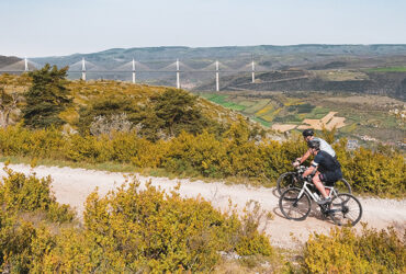 Deux cyclistes gravel roulent sur piste avec au loin le Viaduc de Millau et ses vallées dorées