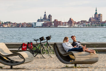 Un couple de cycliste se repose sur une plage en face d'une ville aux briques rouges