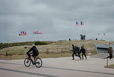 Un cycliste roule le long de la plage et devant une statue de soldats